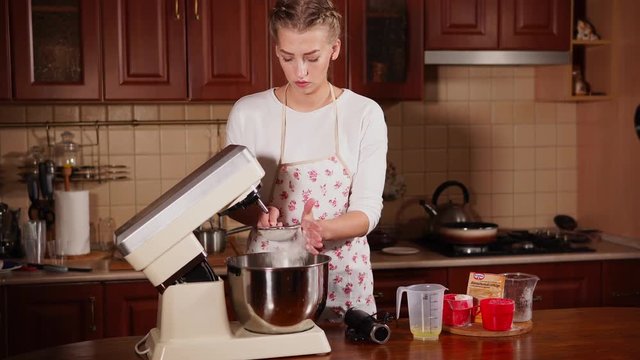Lovely blonde female cook is getting ready for baking. She is adding to sieves of flour in the bowl of mixer.
