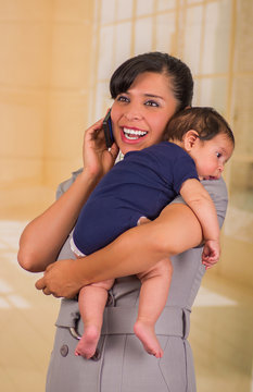 Young mother is holding her little baby and using hger cellphone in a blurred background