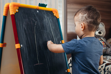 a young three year old boy drawing with chalk on a blackboard on an easel, at home in my room