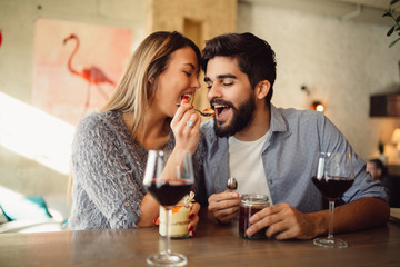 Beautiful couple having romantic moments in cafe, girl feeding her boyfriend with cake in cafe, drinking wine and celebrating Valentine's day.