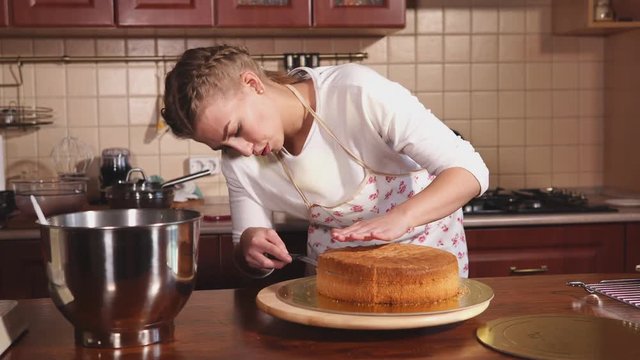 a young woman who looks like a chef cuts cakes for making a layered cake with cream cream, the lady is regulating a knife dough on a stand that is on the table