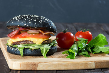 Large burger with a black buns/ Large burger with a black buns and bacon on a cutting board with a wooden countertop. Tomatoes and lettuce leaves.