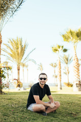 Handsome bearded man in sunglasses sitting on the grass under palms on summer vocation.