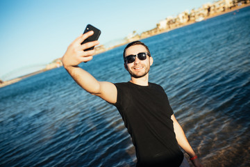 happy smiling young man in sunglasses and hat taking selfie on beach
