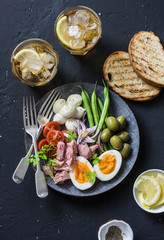 Appetizer table - plate of canned tuna, green beans, mozzarella cheese, tomatoes, boiled egg, olives, grilled bread and two glasses of white wine on dark background, top view