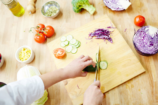 Hands of chef cutting fresh cucumber on wooden board at workplace