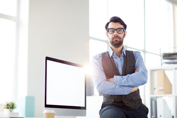 Serious office worker with his arms crossed on chest sitting on his desk and looking at camera