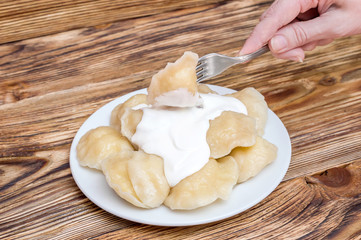 Hand holding boiled varenyky on fork and plate with vareniki on wooden table.