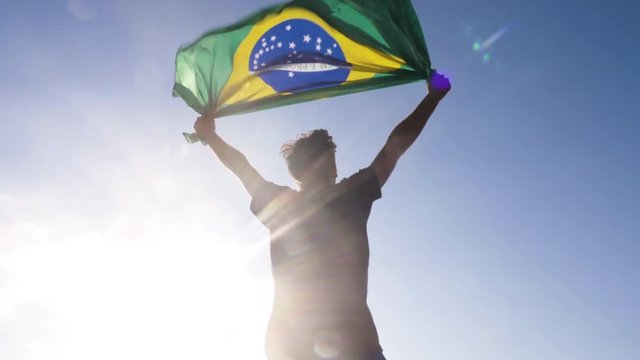 Young man holding brazilian national flag to the sky with two hands at the beach at sunset brazil