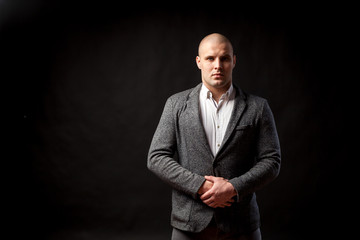 A young bald man in a white shirt, gray suit confidently looks at the camera and holds his hands on his jacket on a black isolated background