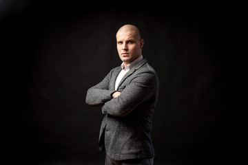 A young bald man in a white shirt, gray suit confidently looks at the camera and holds his arms crossed on his chest on a black isolated background