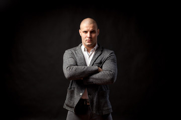 A young bald man in a white shirt, gray suit confidently looks at the camera and holds his arms crossed on his chest on a black isolated background