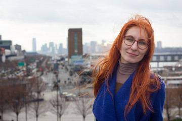 Cute red-haired girl in a blue coat and glasses standing on the background of the big city, her hair fluttering in the wind