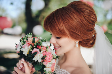 Naklejka premium A young bride in a white lace dress is sniffing her wedding bouquet that is carefully held by her hands. Red-haired bride.