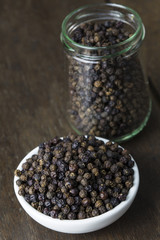 Pile black pepper on white bowl and glass jar on old wooden table.