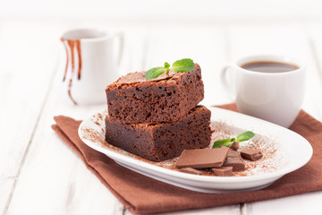 Fototapeta na wymiar Chocolate brownie square pieces in stack on white plate decorated with mint leaves and cocoa powder on white vintage wooden background. American traditional delicious dessert. Close up photography