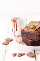 Chocolate brownie square pieces in stack on white plate decorated with mint leaves and cocoa powder on white vintage wooden background. American traditional delicious dessert. Close up photography