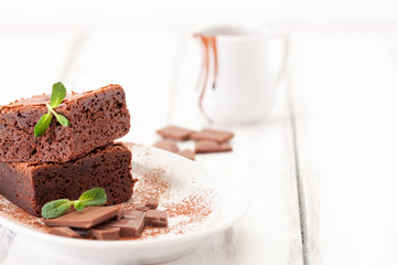 Chocolate brownie square pieces in stack on white plate decorated with mint leaves and cocoa powder on white vintage wooden background. American traditional delicious dessert. Close up photography