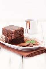 Chocolate brownie square pieces in stack on white plate decorated with mint leaves and cocoa powder on white vintage wooden background. American traditional delicious dessert. Close up photography