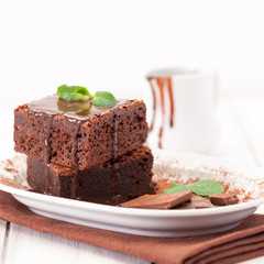 Chocolate brownie square pieces in stack on white plate decorated with mint leaves and cocoa powder on white vintage wooden background. American traditional delicious dessert. Close up photography