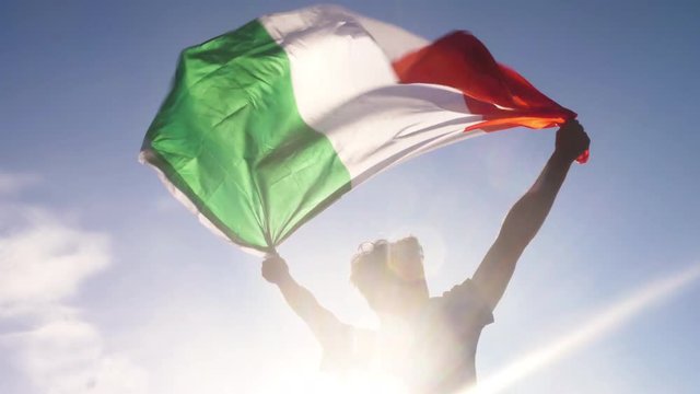 Young man holding italian national flag to the sky with two hands at the beach at sunset italy