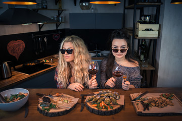Two young women holds glasses of wine and sits together in modern home kitchen at the bar counter. Attractive people sitting at the table with food. Girlfriends enjoying evening together