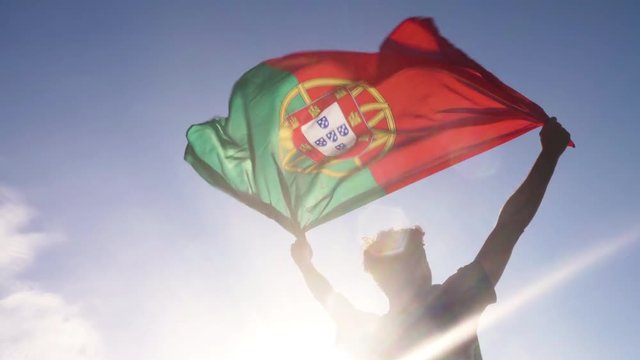 Young man holding portuguese national flag to the sky with two hands at the beach at sunset portugal