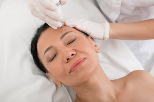 Top view of glad mature woman relaxing during laser procedure in beauty salon. Her eyes are closed. She is lying on the couch