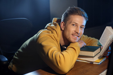 Portrait of cheerful bearded young male looking through book. Different tomes locating on table. Knowledge and hobby concept