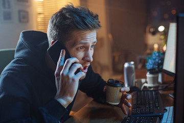 Portrait of serene bearded man telling by mobile while drinking mug of hot beverage. He having job with gadgets at home. Occupation and appliance concept