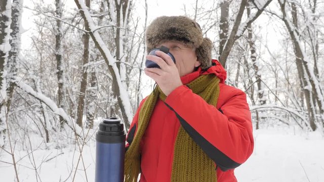 Ederly European man drinking tea from a thermos bottle in a snow-covered forest. Thick dense thicket of trees and roots in in the snow-covered forest. Hike and travel concept 60 fps