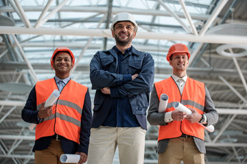 Professional team. Low angle portrait of joyful young builders are standing together and looking ahead with smile while holding blueprints. They are wearing safety-helmets