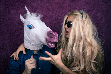 Portrait of happy unicorn in the arms of freaky young woman in sunglasses and kitty ears on the purple background. Expressive blond with strange guy. Unusual people shows emotions. © Sergey_T