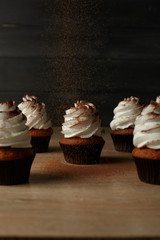 Cupcakes with white cream on which cocoa powder is poured on a wooden table with a dark background