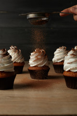 Cupcakes with white cream on which cocoa powder is poured on a wooden table with a dark background