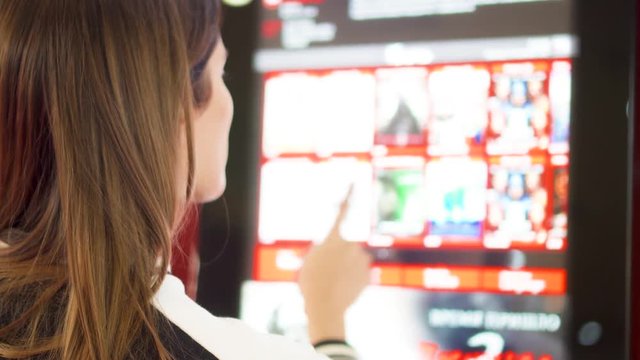 Teenager choosing movie and buying ticket from vending machine at movie theater at mall. Young woman making gestures by touching screen