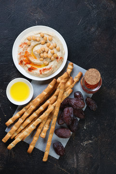 Above view of salted breadsticks with hummus and date fruits, dark brown stone surface, vertical shot