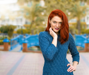  Red-haired girl with freckles posing in the park. Woman in blue woolen beach smiling at the camera.