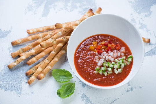 Bowl of gazpacho tomato soup with grissini and fresh green basil on a white concrete background, horizontal shot