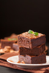 Chocolate brownie square pieces in stack on white plate with walnuts, decorated with mint leaves and cocoa on black background. Delicious dessert. Dark mood. Close up photography. Selective focus