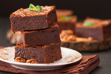 Chocolate brownie square pieces in stack on white plate with walnuts, decorated with mint leaves and cocoa on black background. Delicious dessert. Dark mood. Close up photography. Selective focus