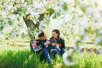 Joyful man, woman rest on nature hug, play, kiss with little cute child baby boy. Mother, father, little kid son sit under white flower tree. Family sun day 15 of may, love, parents, children concept.