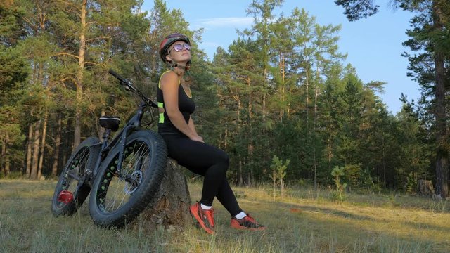 Fat bike also called fatbike or fat-tire bike in summer riding in the forest. Beautiful girl and her bicycle in the forest. She sits on the stump and rests from the trip.