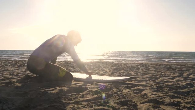 Young man surfer in wetsuit rubbing top coat wax paraffin on surf board on sandy beach at sunset preparing