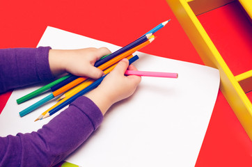 The child's hands are painted with colored pencils on a white sheet of paper on a red table