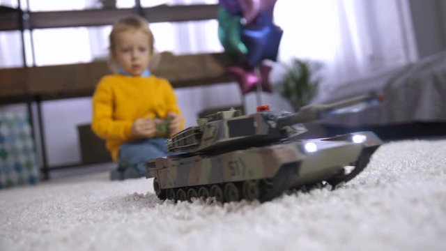 Cute blond toddler boy playing with his new present, remote controlled military tank on the carpet in bedroom. Smart little boy enjoying his birthday present. Rack focus