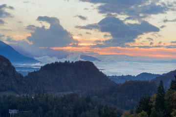 There are a lot of clouds in the distance as the sun rises. There are mountains and trees in the foreground and the sun rise in the background. Taken at Lake Bled in Slovenia.