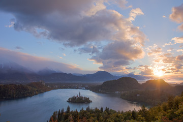 The sun bursts through the clouds overlooking Lake Bled Island in Slovenia. Golden hour is in full effect as the landscape changes color dramatically.