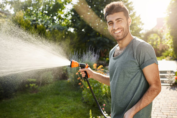 Outside portrait of young attractive caucasian gardener with beard and stylish hairstyle in blue t-shirt smiling in camera, watering plants with garden tool, productive summer morning.