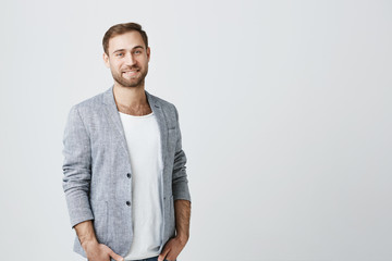 Smiling attractive handsome guy with beard in trendy gray jacket over t-shirt posing against gray...